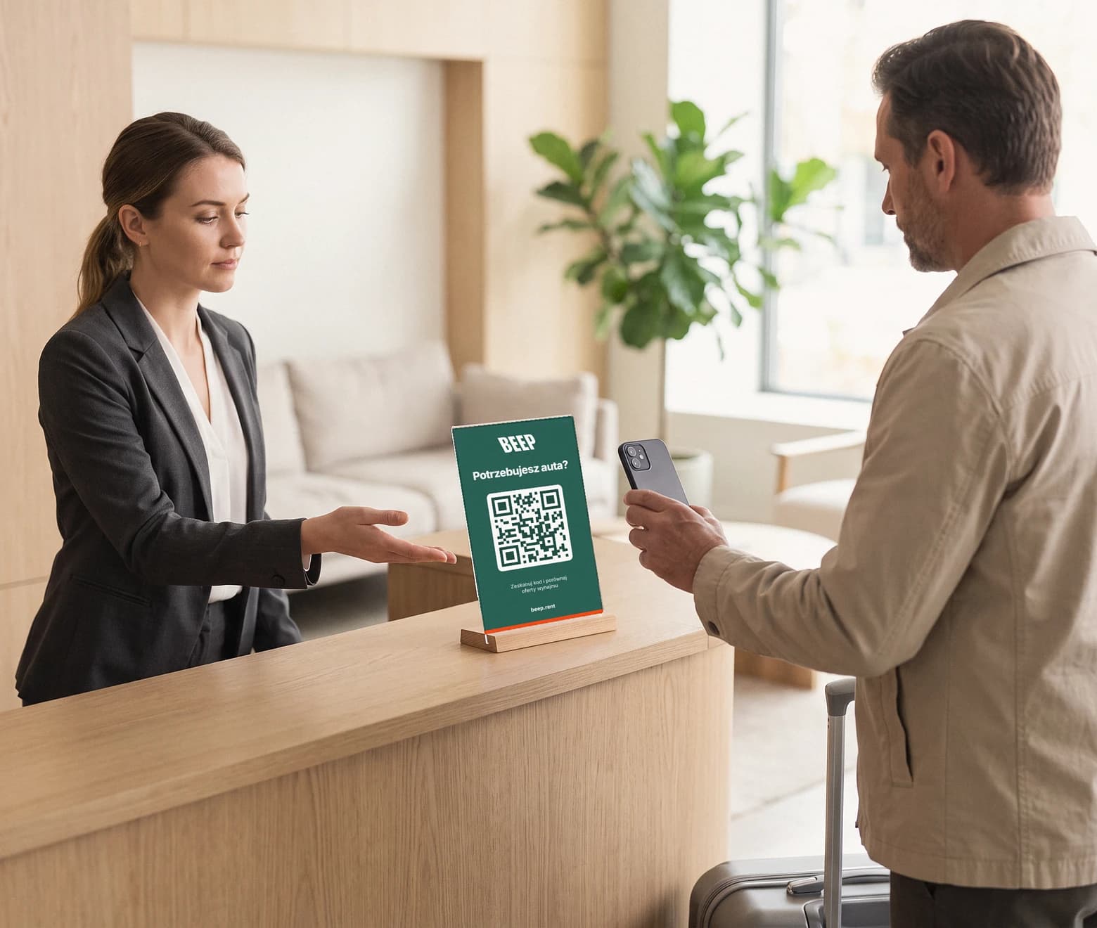 Recepcja hotelu: gość skanuje telefonem kod QR BEEP ze stojaka na ladzie, recepcjonistka wskazuje stojak.